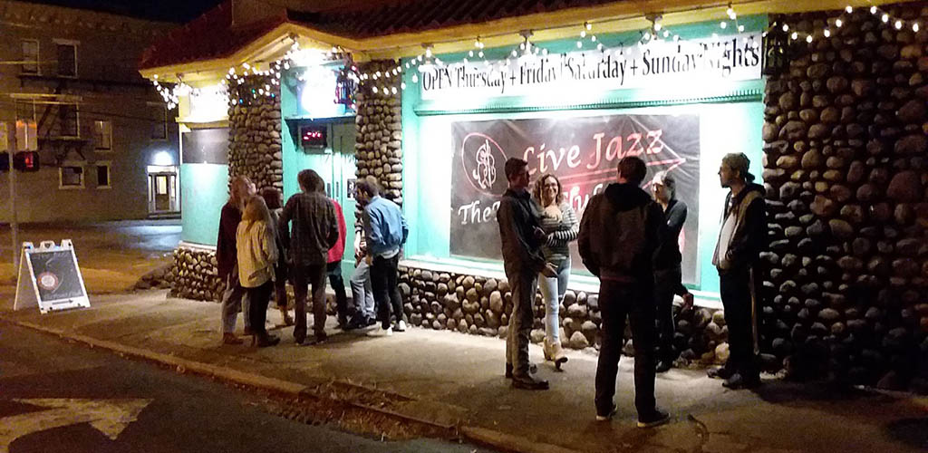 Two small groups of people hanging out in front of a brightly-lit bar